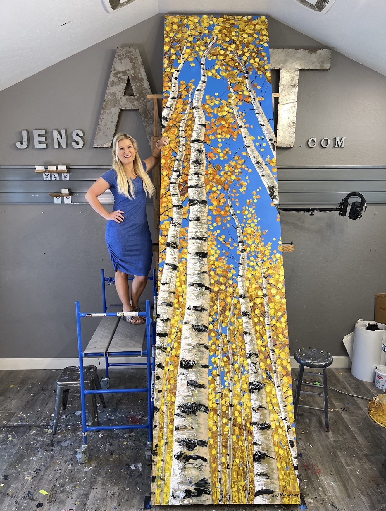 Jennifer Vranes on studio scaffolding beside a tall autumn aspen painting at JensArt